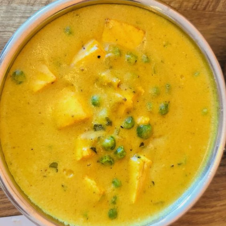 Close-up of a vibrant yellow Indian curry with paneer cubes and green peas in a metal bowl.