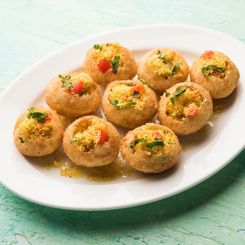 A white plate holds several golden, crispy Indian pani puri, filled with savory toppings.