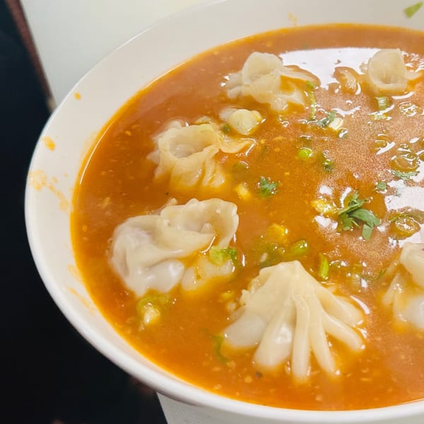 A white bowl filled with momo dumplings in a rich orange Indian curry sauce, garnished with herbs.